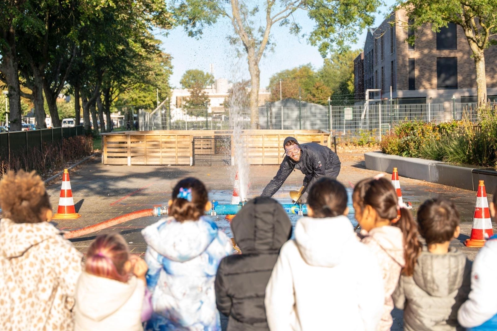 Kinderen kijken toe bij een buitendemonstratie waar water uit waterpijp spuit, afgeschermd met veiligheidspionnen.