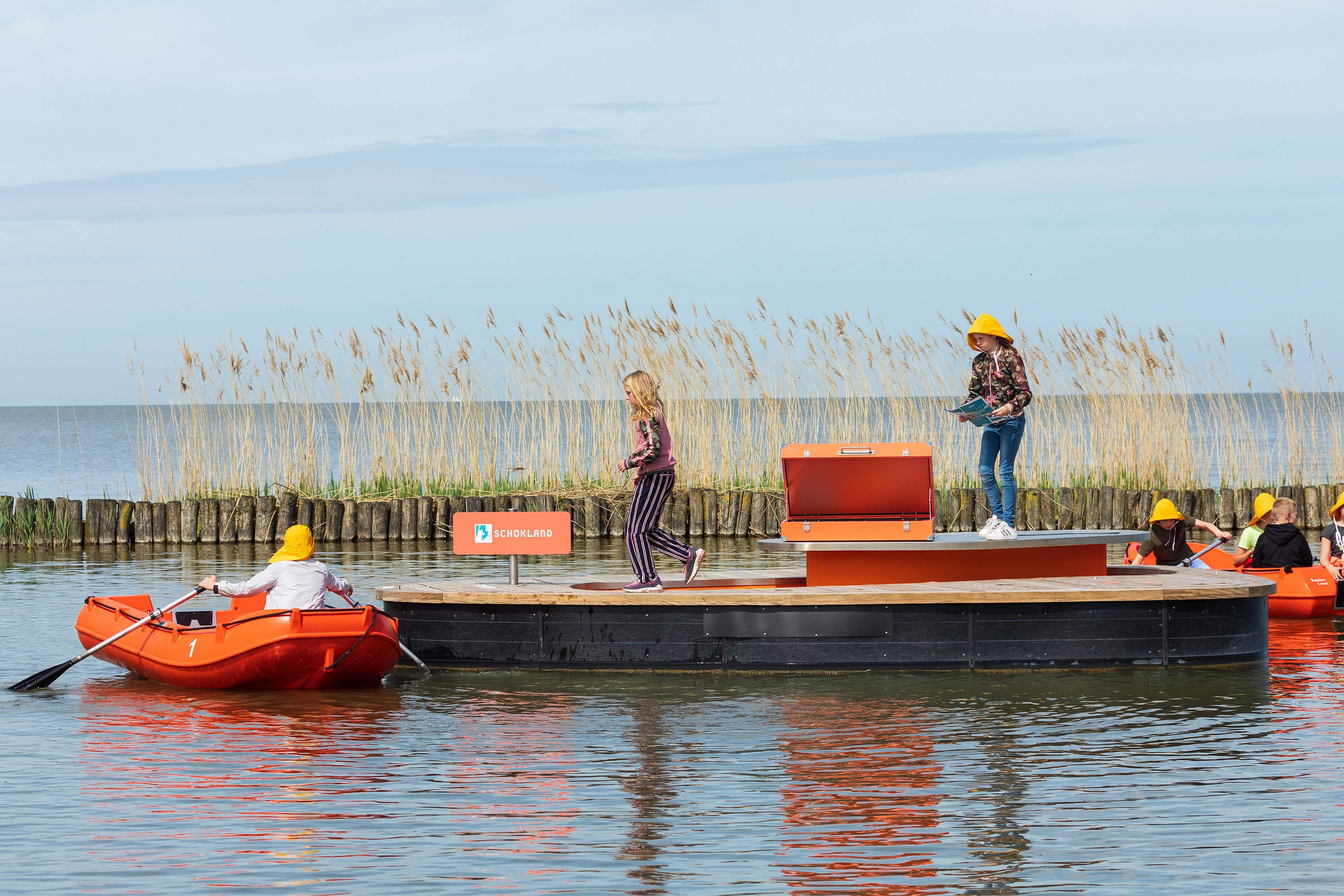 Roeiboten naderen een vlot met open kist en het bord “Schokland”, onderdeel van een educatieve waterexpeditie.