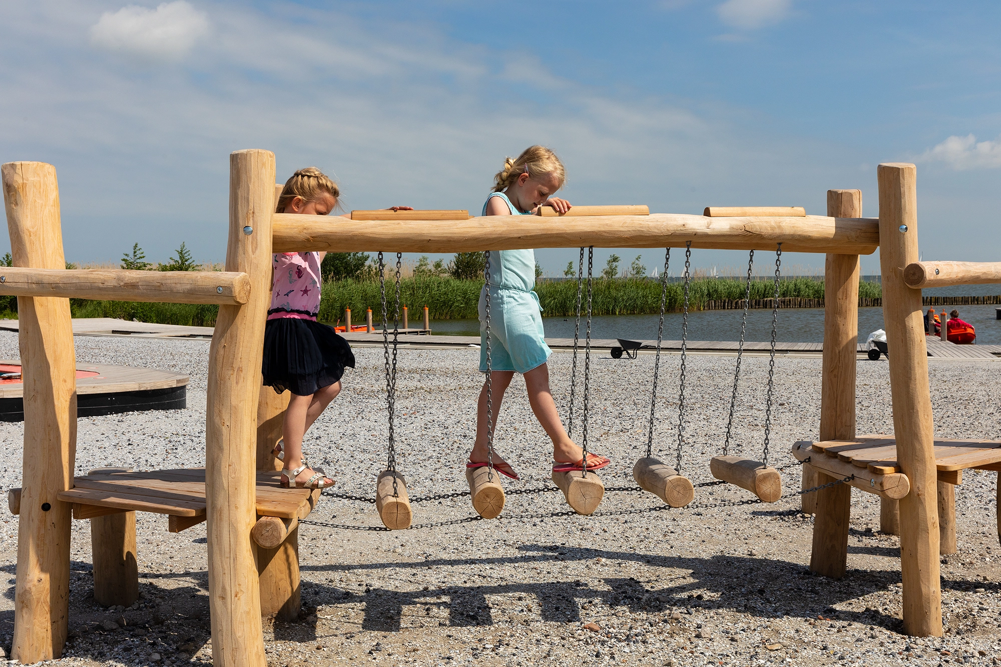 Twee kinderen balanceren op een houten loopbrug bij een buitenspeelplek.