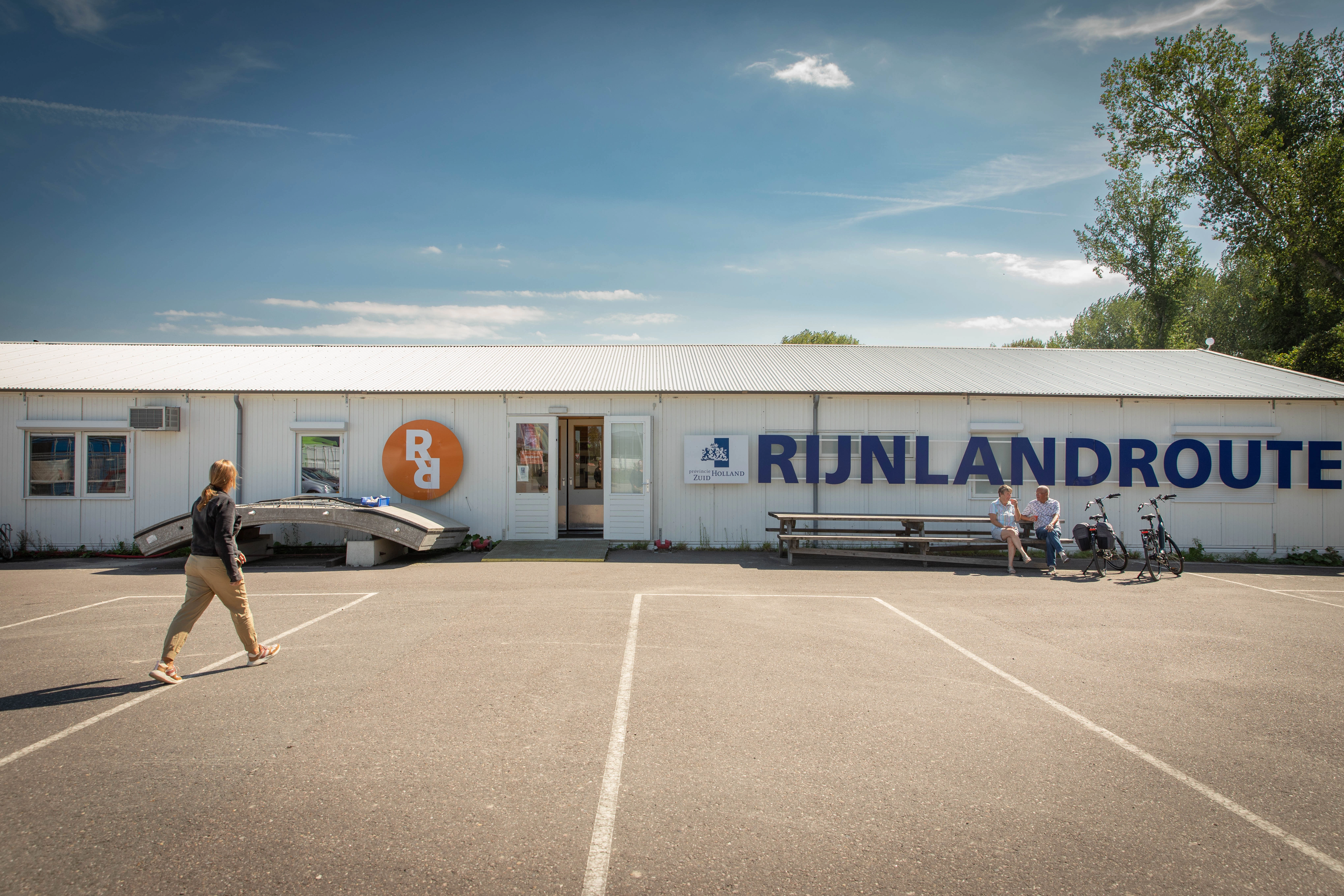 Buitenkant van het informatiecentrum, waarbij iemand naar het infocentrum toe loopt. Op de buitenkant staat in grote letters Rijnlandroute